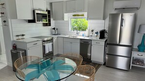 A modern kitchen with white cabinetry, stainless steel appliances, a glass dining table with wicker chairs, and a window with a view of greenery. - The Historic Coquina Cottages C is 2 Blocks to the Beach, 5th Avenue and 3rd St. (Naples)