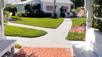 A white gazebo with a gravel pathway leading to a house with a metal roof and pink shutters.