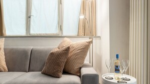 Una sala moderna con un sofá gris, cojines beige, una mesita de noche blanca con una botella y vasos, y una ventana con cortinas.