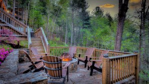 Una terraza de madera con sillas Adirondack y un pozo de fuego de piedra rodeado de bosque.