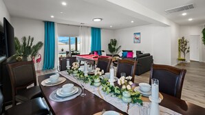 Ett matområde med ett långt bord dukat för sex personer, omgivet av stolar. I bakgrunden finns ett biljardbord, och rummet har en modern design med vita väggar och tak.
