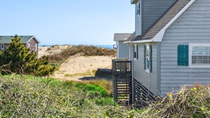 A coastal house with a wooden deck, a sandy beach in the background, and a clear sky.