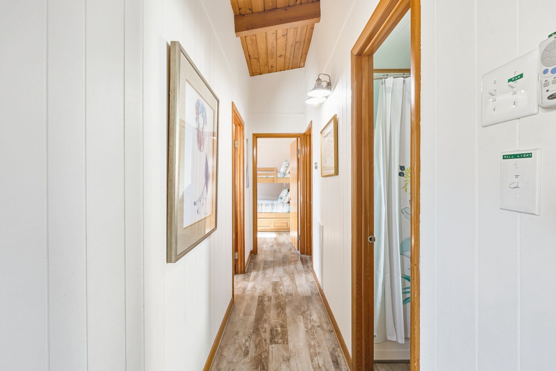A hallway with wooden flooring, white walls, and framed artwork. There is a door leading to a room with a bunk bed.
