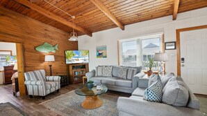 A cozy living room with a wooden ceiling, a grey sofa, a striped armchair, a glass coffee table, and a TV on a wooden cabinet.