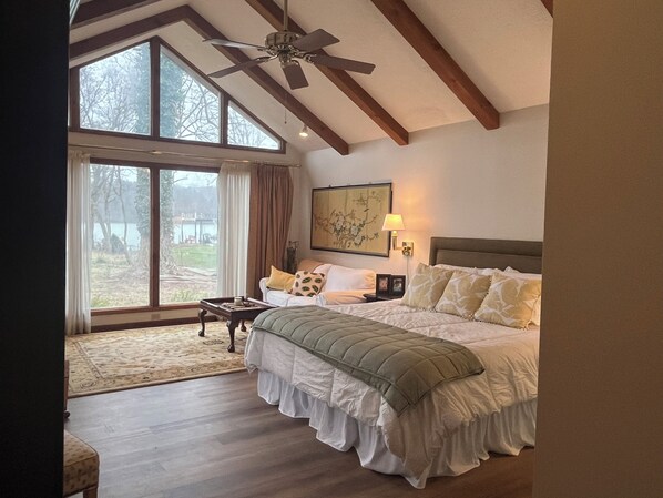 Ein geräumiges Schlafzimmer mit einem großen Bett, einem Deckenventilator, Seeblick durch große Fenster und Holzboden.
