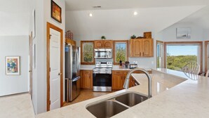 A modern kitchen with stainless steel appliances, wooden cabinets, a large island with a sink, and a view of greenery outside. - Despacito: Pool, Hot Tub, Walk to Beach OBX (Corolla)