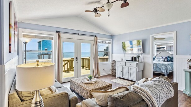 Un salón espacioso con vista al mar, un balcón con terraza de madera, un aparador blanco y una cama con colcha azul y blanca.