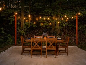 An outdoor dining area with a wooden table and chairs, surrounded by trees and string lights. - Cozy Home Near Downtown | Newly Added Amenities! | (Cedar)