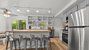 A modern kitchen with a central island, bar stools, stainless steel appliances, and built-in shelving. - Cozy Home Near Downtown | Newly Added Amenities! | Labor Day Open! (Cedar)