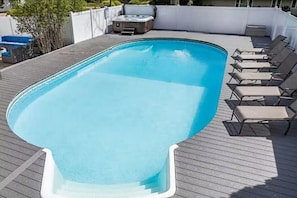 A rectangular swimming pool with a wooden deck, surrounded by lounge chairs and a hot tub.