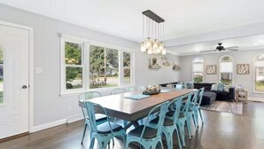 A spacious dining area with a long table and chairs, large windows, and a ceiling fan.