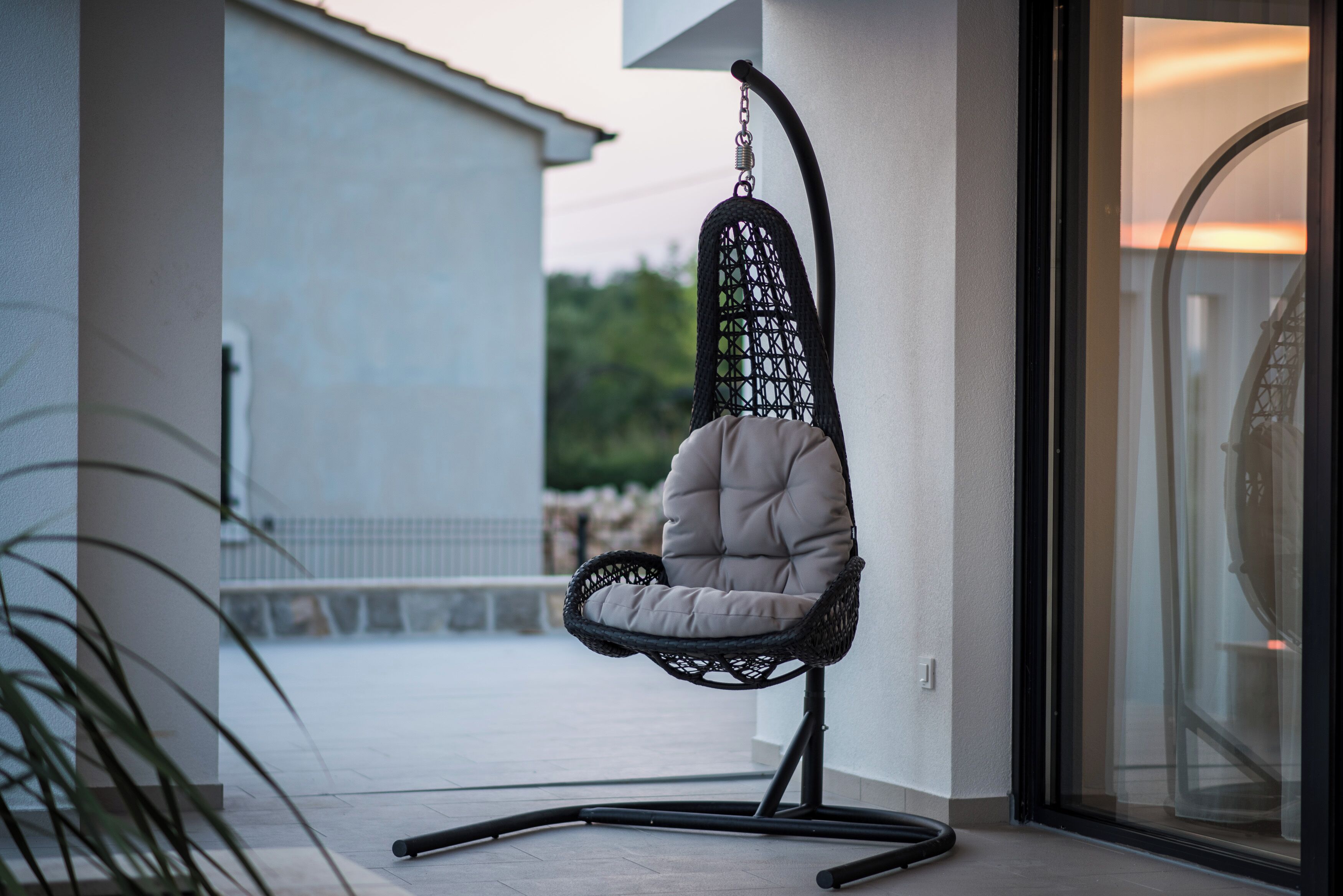 A hanging chair with a cushioned seat and a woven backrest, positioned on a balcony with a view of a building and greenery in the background.