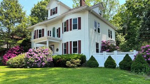 A two-story house with a white exterior and red shutters, surrounded by well-maintained gardens and a white picket fence.