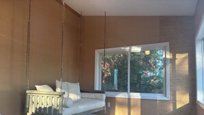 A room with a wooden floor, a white sofa with cushions, and a large window with a view of trees and a fence.