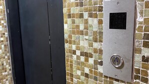Et metallisk betjeningspanel til elevator med en knap med en pil og en lille, rund knap, foran en væg af mosaikfliser.