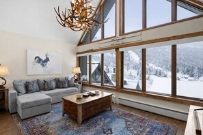 A cozy living room with a gray sofa, patterned cushions, a wooden coffee table, and a large window offering a snowy mountain view.