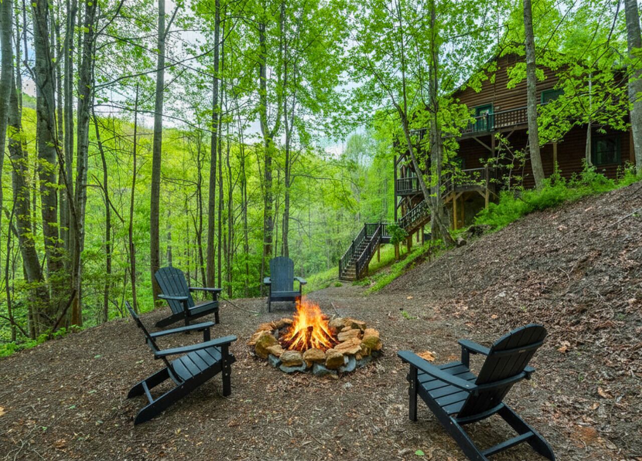 Ein bewaldetes Gebiet mit einer Feuerstelle, umgeben von Adirondack-Stühlen, eine Holzhütte mit Balkon und eine Treppe, die den Hügel hinaufführt.