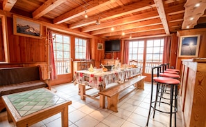 A wooden cabin interior with a dining table, benches, a bar area, and large windows.