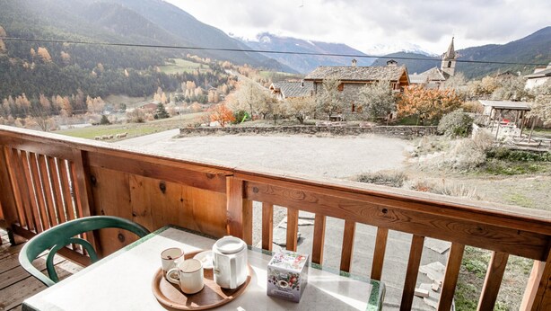 A wooden deck with a table and chairs, a tray with a teapot, cups, and a box, and a view of a village with houses and mountains in the background. - Apartment 'Les Mélèzes' with Mountain View, Balcony and Wi-Fi (SOLLIERES SARDIERES)