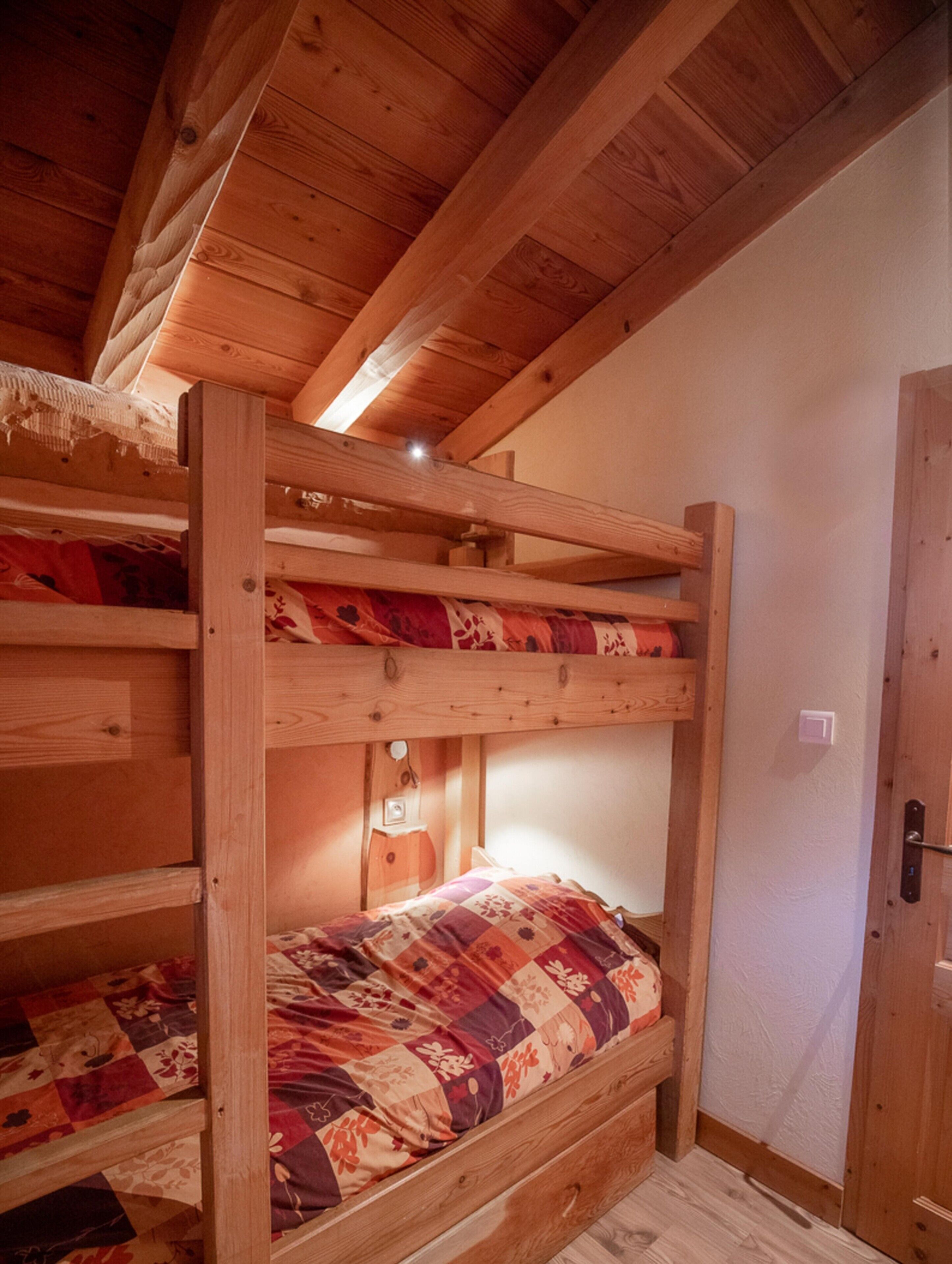 Una cama litera con colcha, vigas de madera, una lámpara fijada a la pared y una puerta.