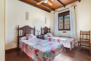 A room with two wooden beds, a wooden chair, a ceiling fan, and a window with curtains.