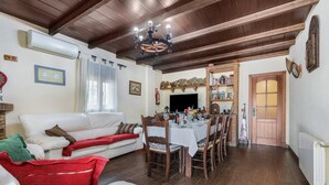 Een woonkamer met een witte bank, eettafel en stoelen, houten balken aan het plafond en een open haard.