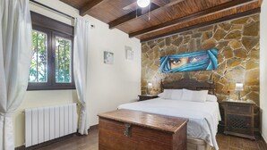 Een slaapkamer met een stenen muur, een bed, een houten kist, een nachtkastje en een raam met gordijnen.