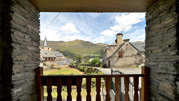 A stone building with a balcony, a church with a spire, and a hill in the background.