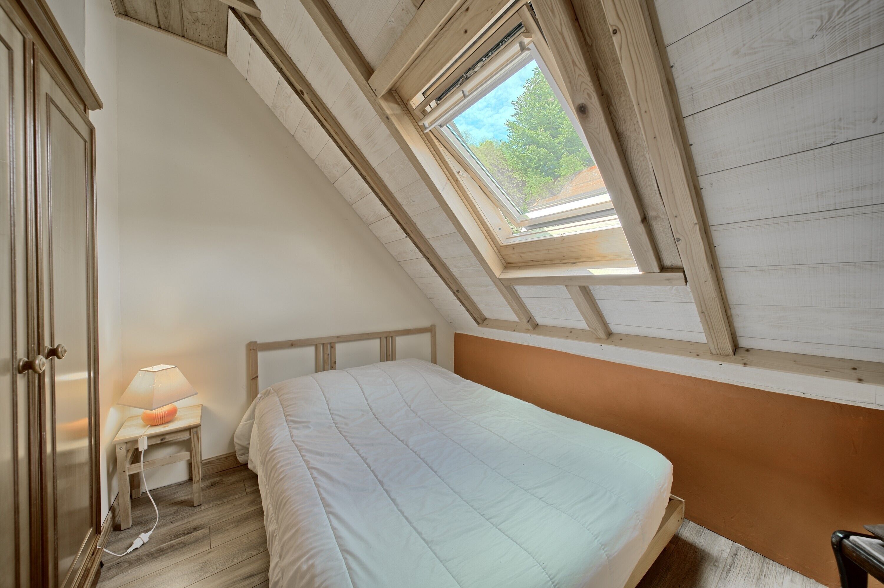 A bedroom with a sloped ceiling, a skylight, a bed, a bedside table with a lamp, and a wardrobe.
