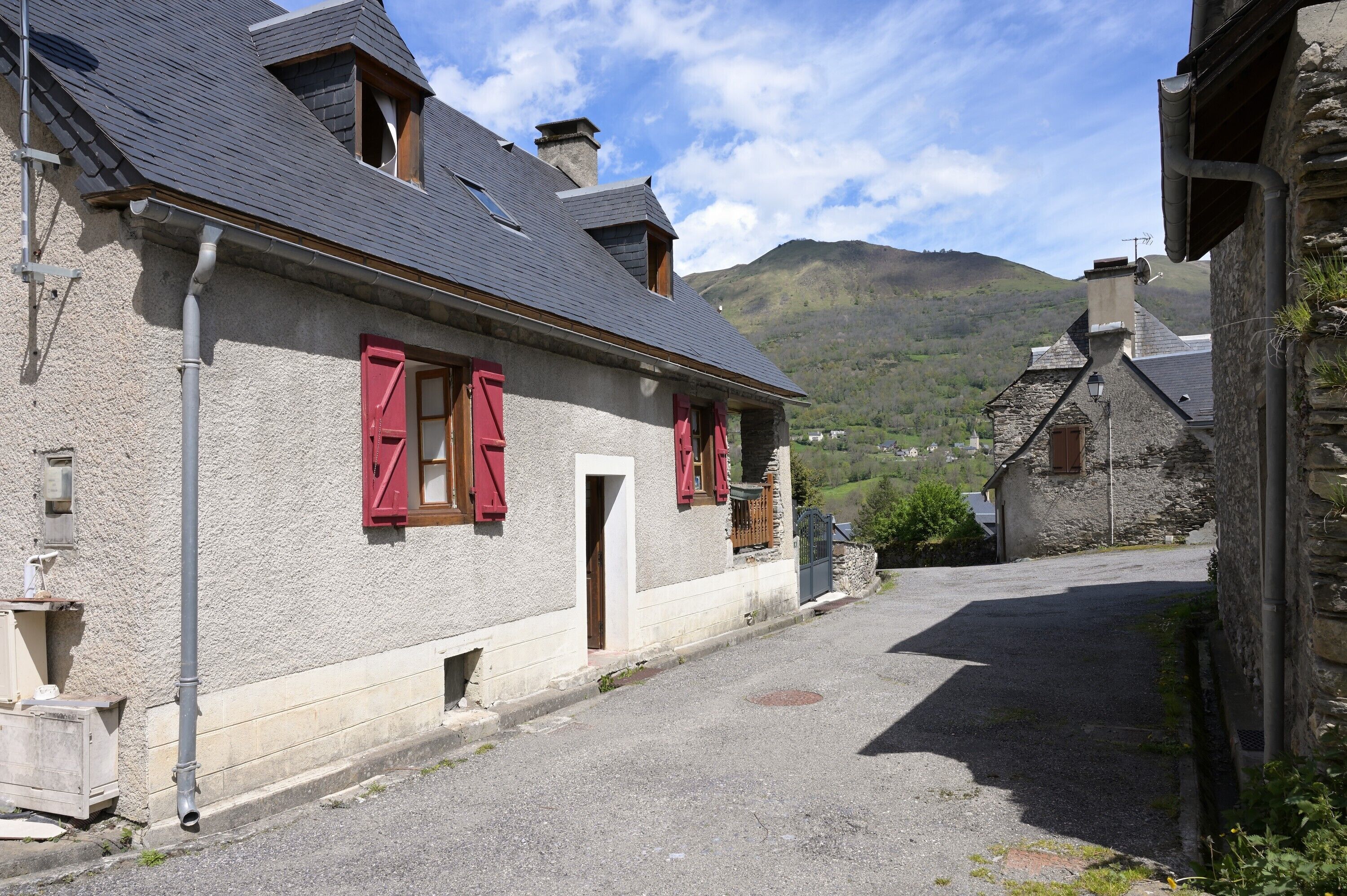 Una calle estrecha con edificios de piedra, contraventanas rojas y una montaña al fondo.