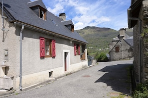 Une rue étroite bordée de bâtiments en pierre, de volets rouges et, au loin, d’une montagne.