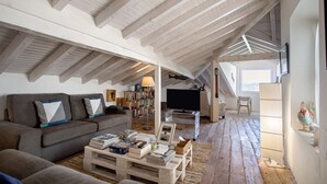 Ein Dachzimmer mit hohen Decken, Holzbalken, einem Sofa, einem Couchtisch mit Büchern, einem Fernseher und einem Fenster mit Blick.