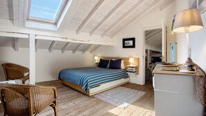 Ein Schlafzimmer mit Dachfenster, einem Bett mit gestreifter Bettdecke, einem Korbstuhl, einem Nachttisch mit Lampe und einem kleinen Schreibtisch ebenfalls mit Lampe.