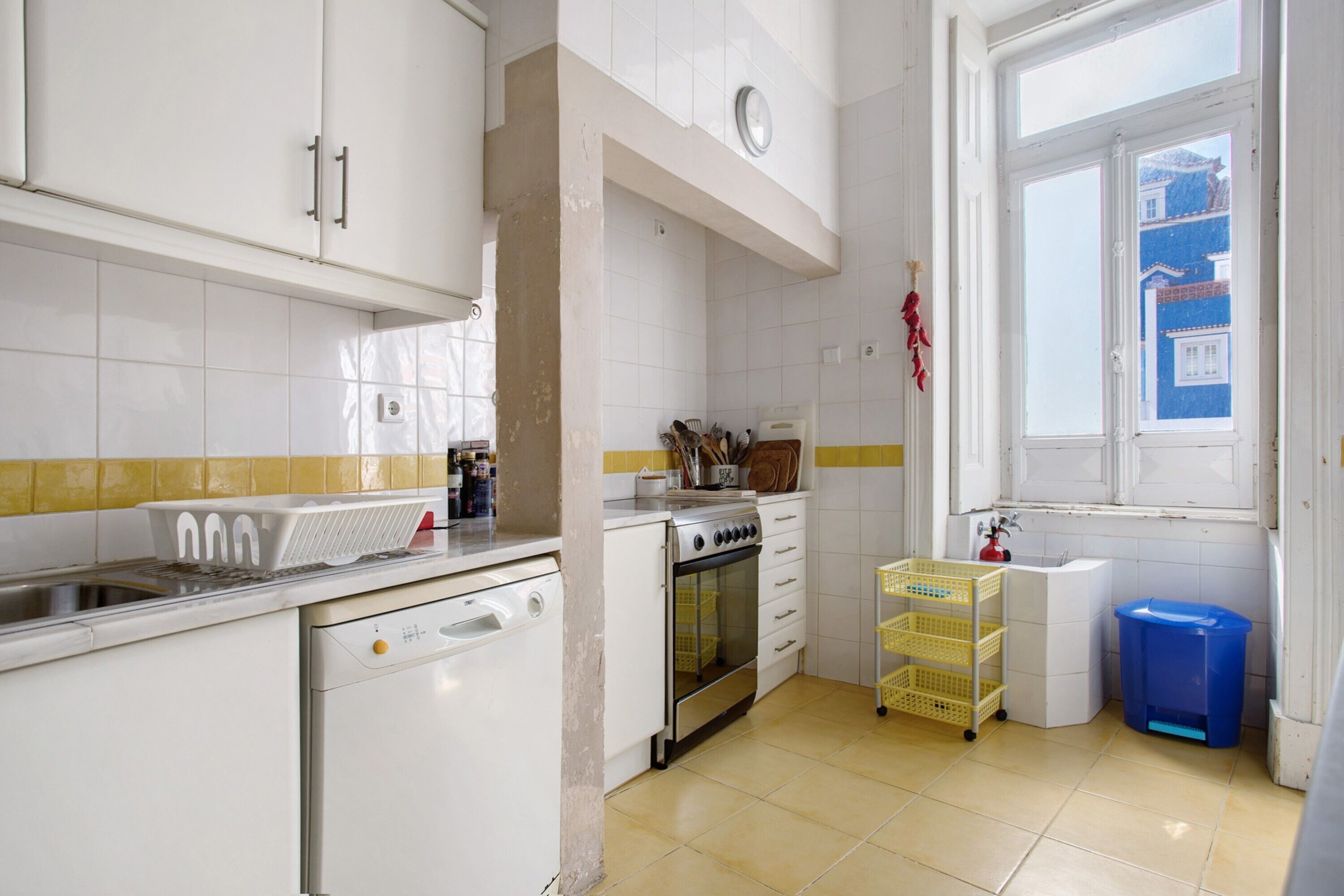Una cocina con armarios blancos, lavavajillas, horno, pileta y ventana con vistas a edificios.