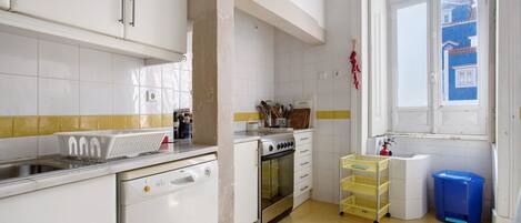 Una cocina con armarios blancos, lavavajillas, horno, pileta y ventana con vistas a edificios.