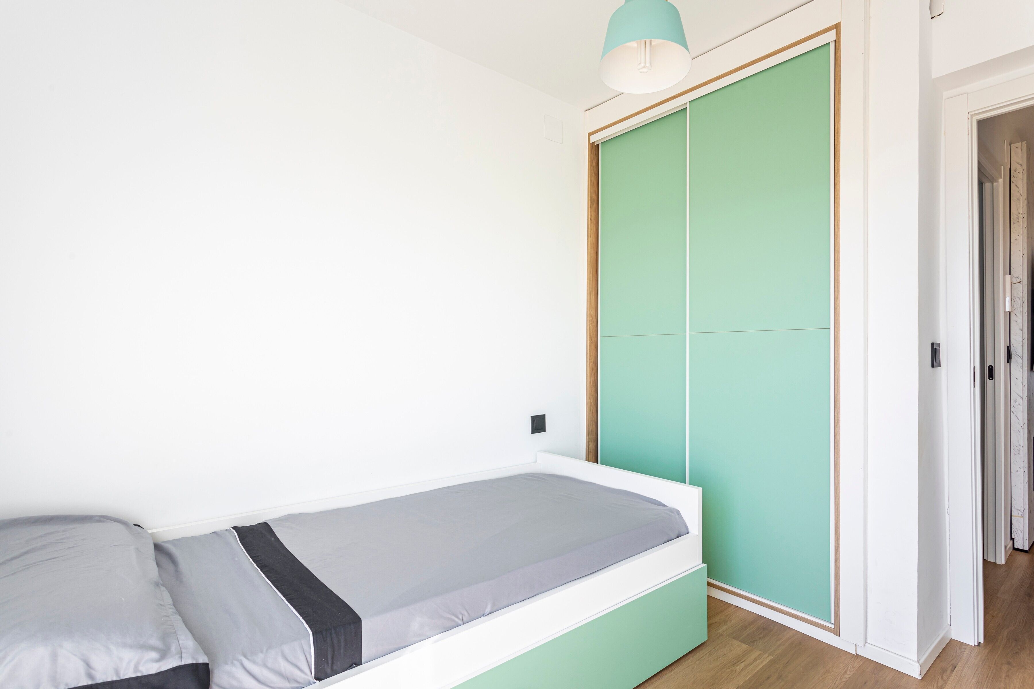 Una habitación pequeña con cama individual, un armario verde y una lámpara de techo blanca.