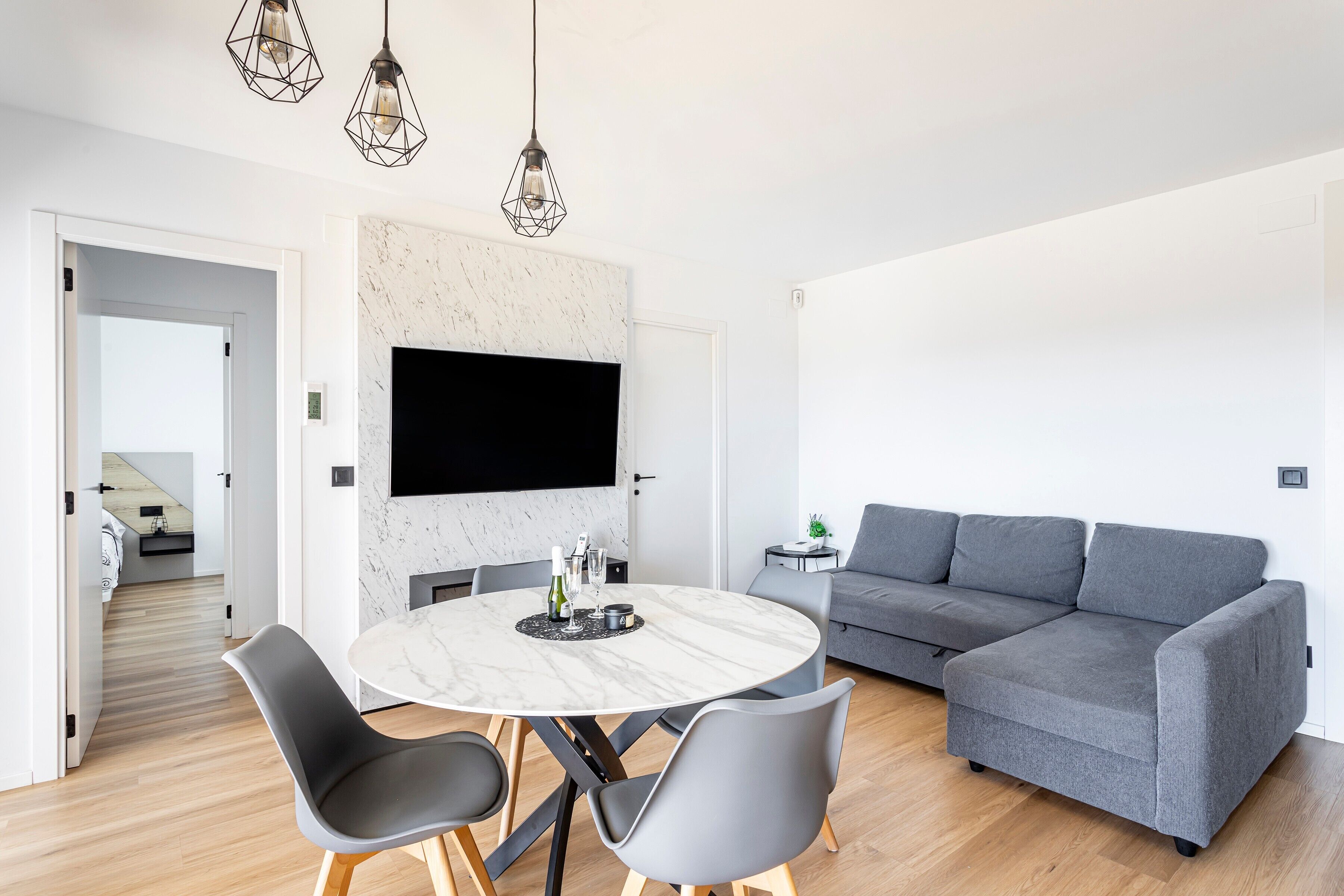 Una sala de estar moderna con una mesa redonda de mármol, un sofá gris y un televisor de pantalla plana montado en una pared blanca.
