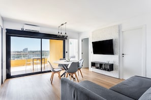 Un salon moderne doté d’un téléviseur à écran plat, d’un coin repas avec une table ronde et des chaises, et d’un balcon offrant une vue sur la ville.
