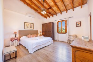A bedroom with a wooden ceiling, a bed with white bedding, a wooden wardrobe, a window with curtains, and a small table with cups.