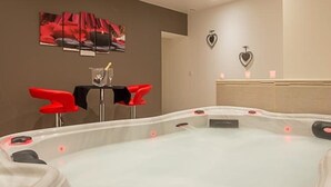 Un cuarto de spa con un hidromasaje, dos sillones rojos, una mesa con una botella y un vaso, y una pared decorada con motivos de corazones.