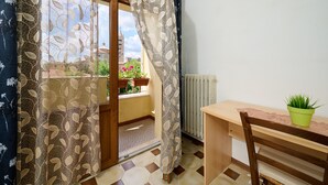 Una habitación con escritorio de madera, una silla, planta en maceta, radiador y vista a un edificio a través de una ventana con cortinas estampadas.