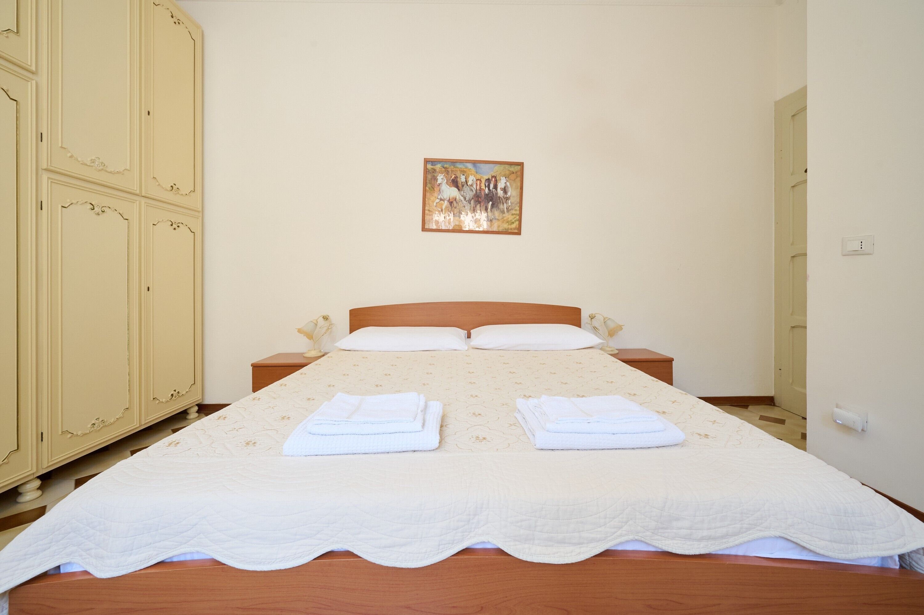 Un lit double avec une couvre-lit matelassé, deux oreillers et des serviettes pliées sur une tête de lit en bois. Un tableau est accroché au mur au-dessus du lit.
