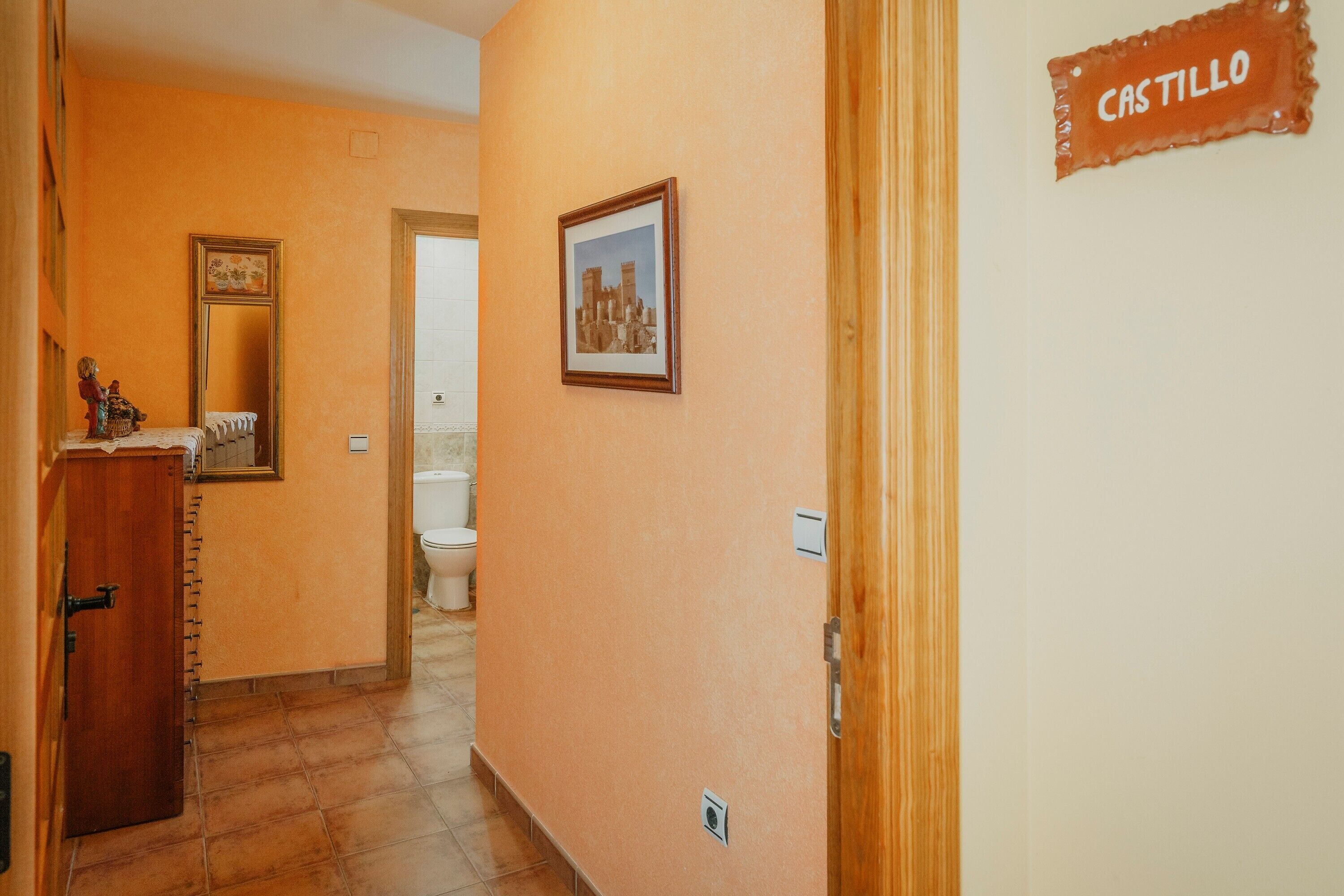 Un pasillo con una puerta de madera que dice 'CASTILLO', un cuadro en la pared y un inodoro a lo lejos.