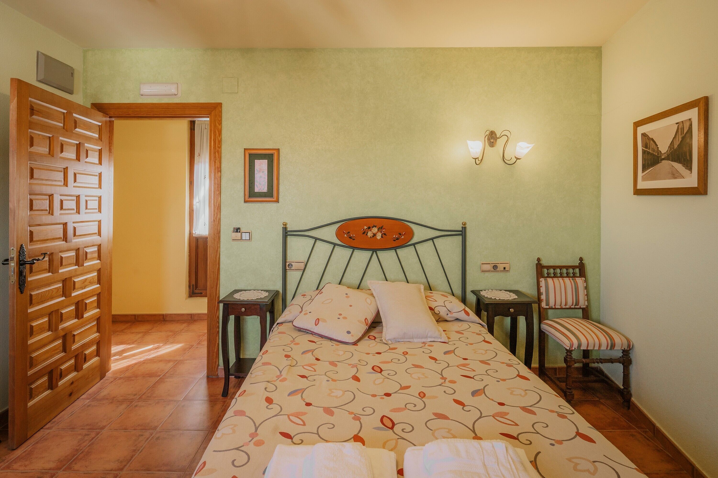 Une chambre à coucher comprenant un lit, une chaise, une table de chevet, une lampe fixée au mur et une porte.