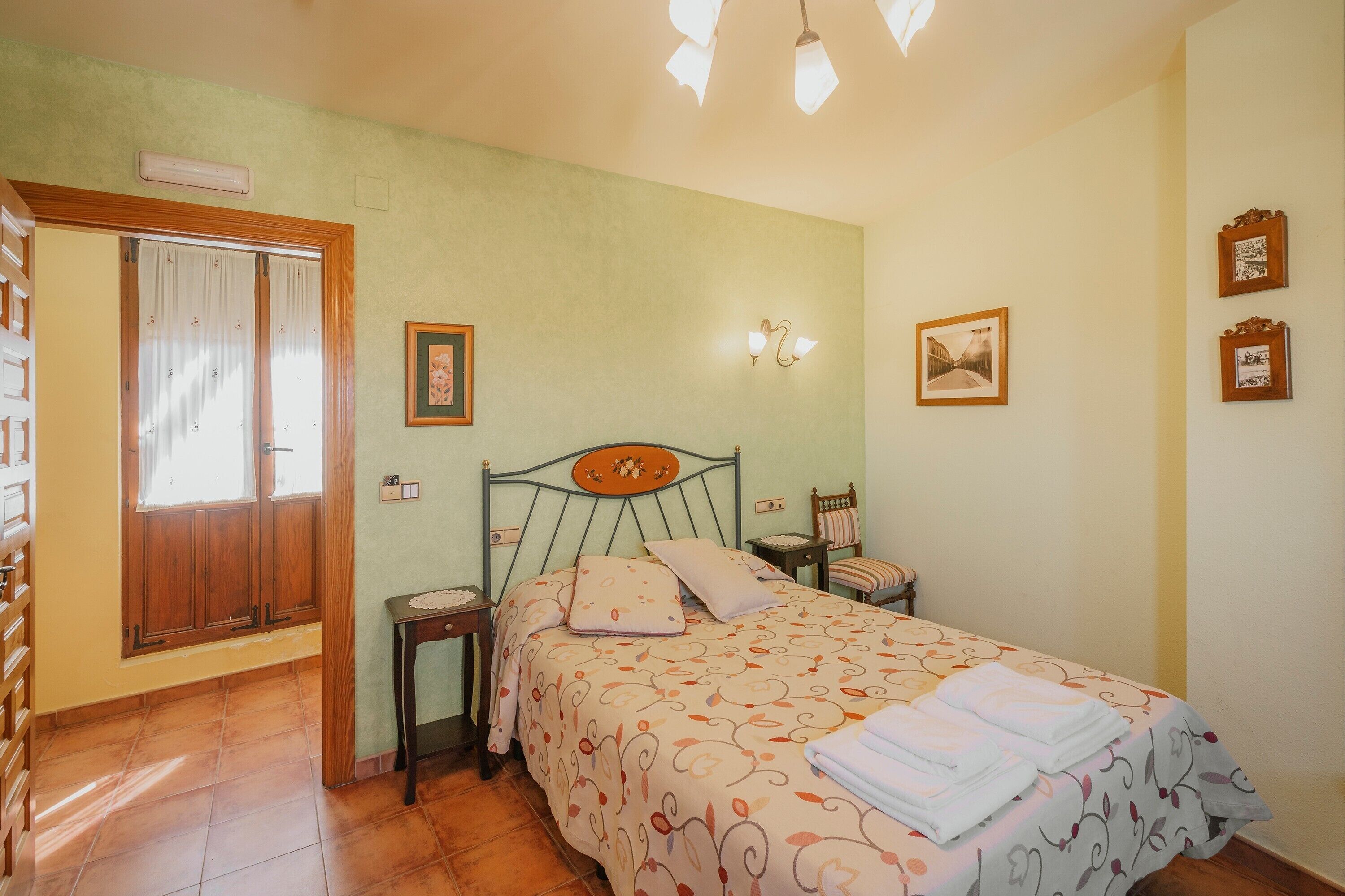 Un dormitorio con cama, mesita de noche, silla, armario de madera y ventana con cortinas.