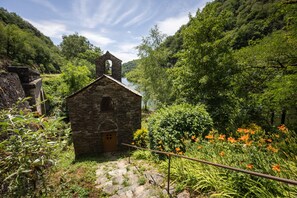 Un bâtiment en pierre doté d’un clocher, entouré d’une végétation luxuriante et bordé par une rivière.