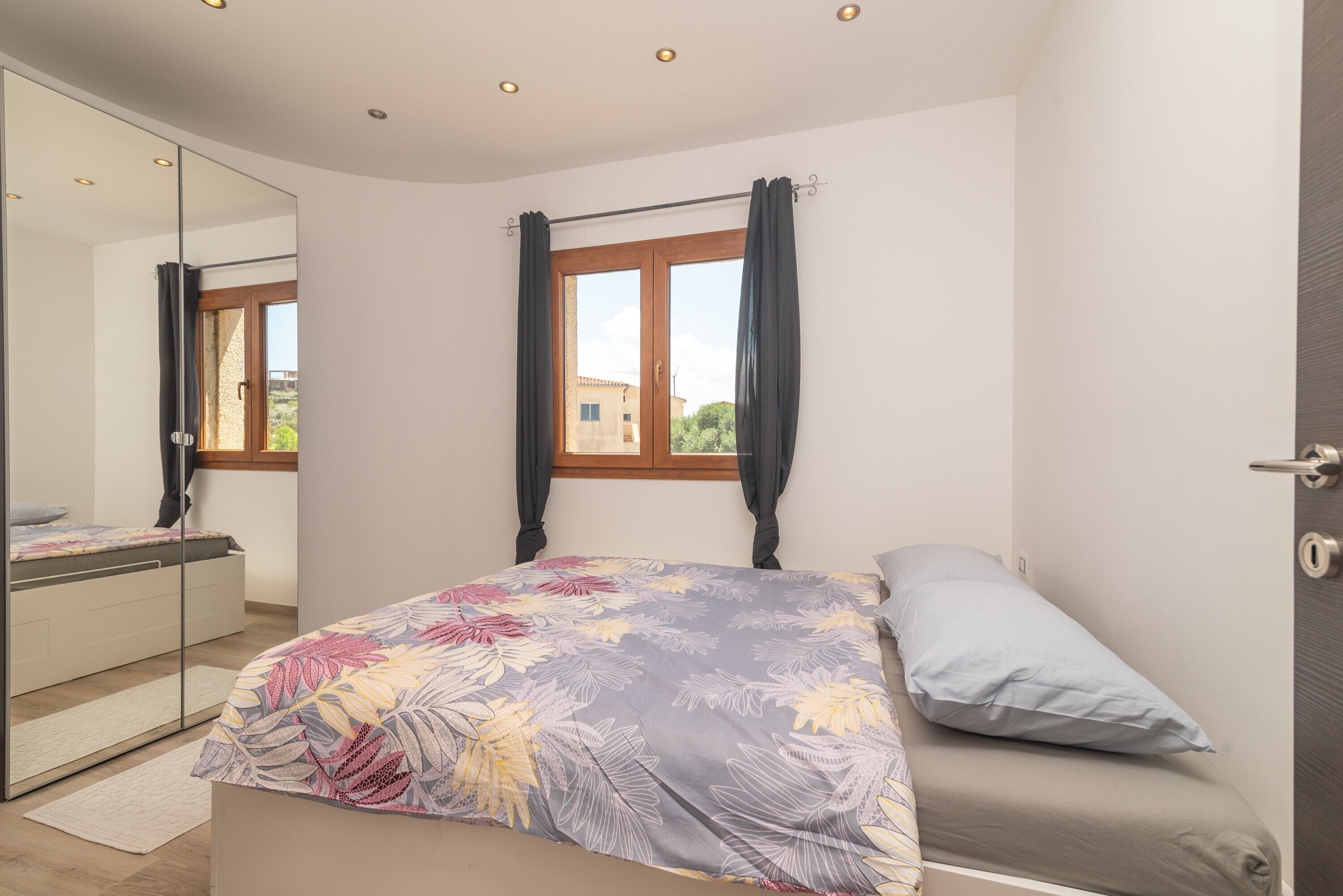 Ein Schlafzimmer mit Bett, Spiegel, Fenster mit Vorhängen und Blick ins Freie.