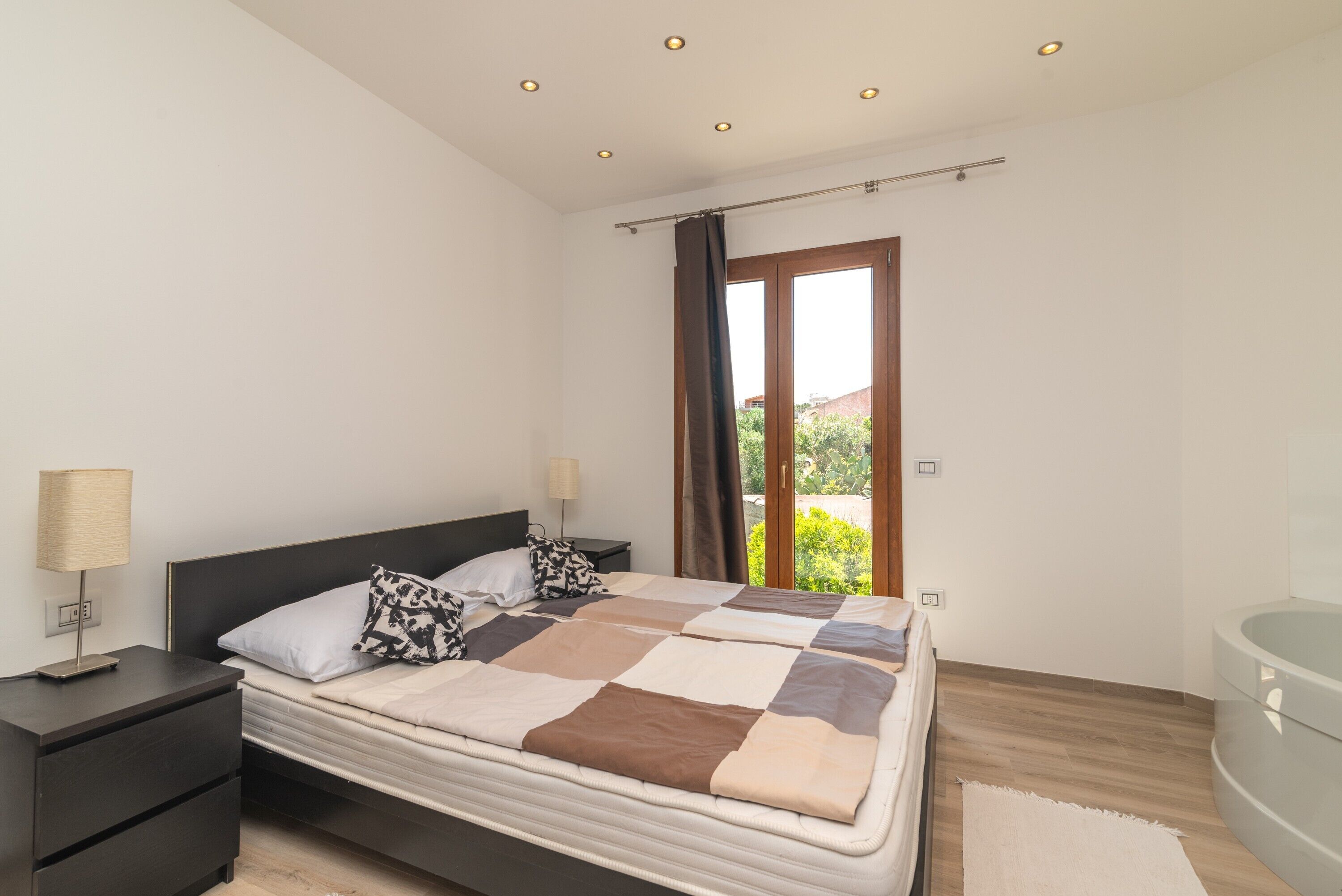 A bedroom with a bed, nightstands, a lamp, a window with curtains, and a view of greenery.