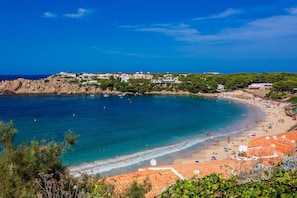 A coastal beach with clear blue waters, a sandy shore, and buildings in the background.