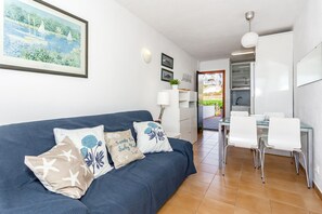 Ein Wohnzimmer mit blauem Sofa, weißen Sesseln, einem Esstisch und einer Küchenzeile mit Schränken und einem Kühlschrank.
